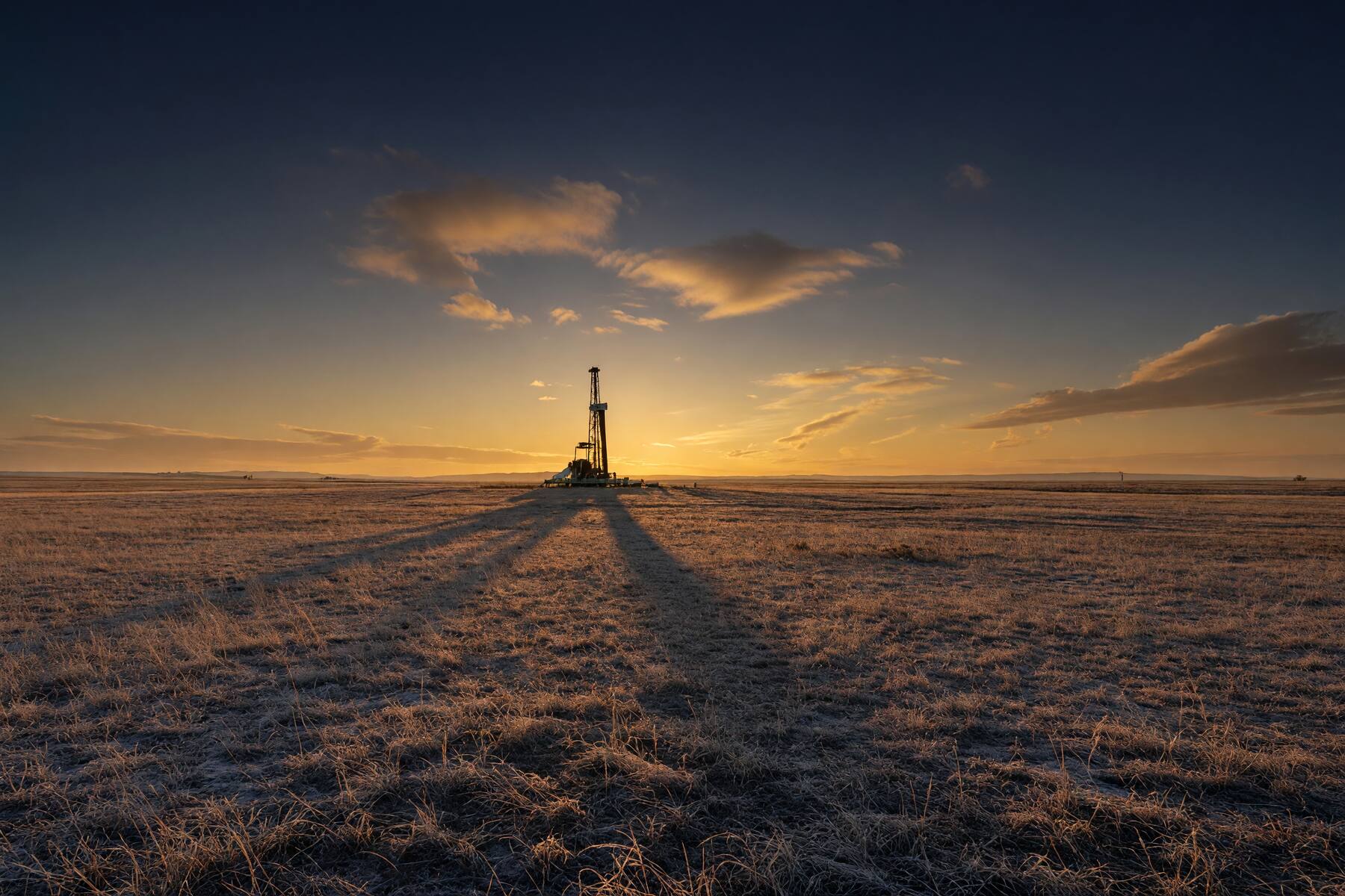 A lone drilling rig silhouetted against a golden-hour sky on a frosted, flat West Texas plain, its long shadow stretching across winter grass.
