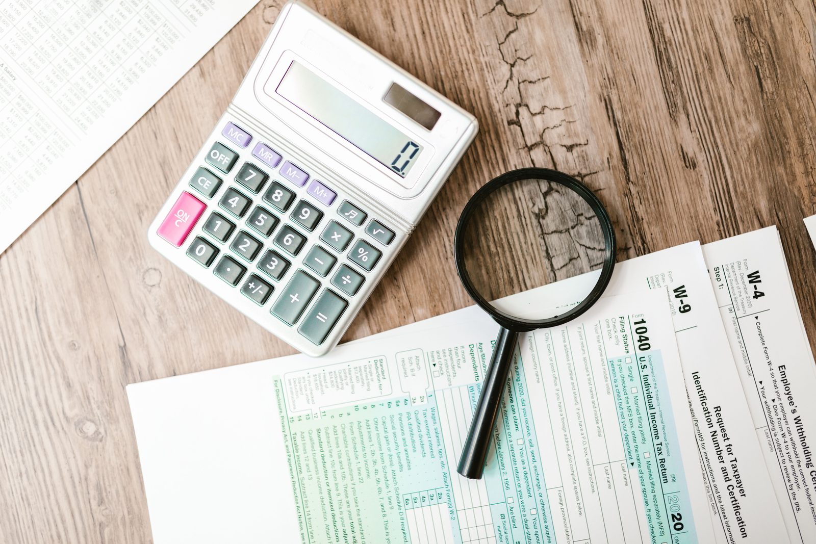 Tax forms laid out with a calculator and a magnifying glass on a wooden surface.
